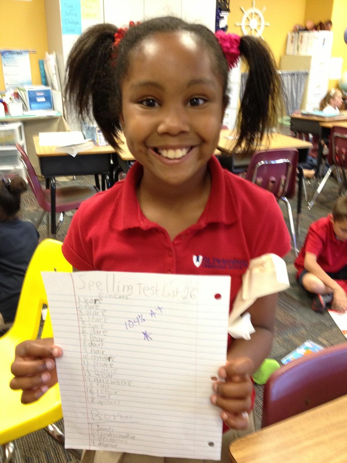 Kimberly's daughter in her classroom holding up a spelling test marked 104% A+
